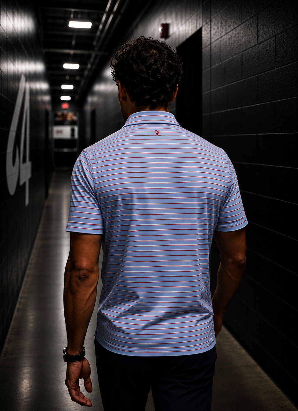 A man viewed from behind walking down a hallway inside of an ice rink  wearing a light blue and white striped polo shirt with thin red stripes.