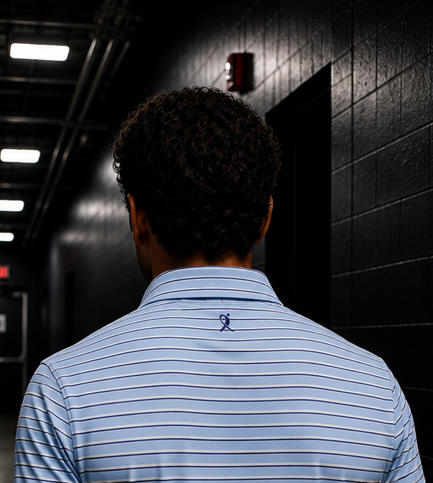 A man viewed from behind walking down a dark hallway in an ice rink, wearing a light blue navy and white striped polo shirt from Nicemitts.