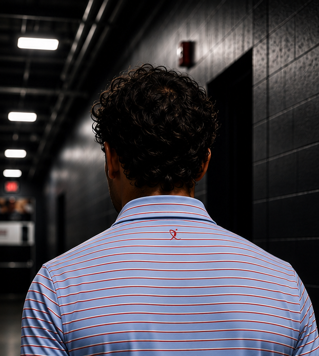 A man viewed from behind walking down a dark hallway in an ice rink, wearing a light blue red and white striped polo shirt from Nicemitts.